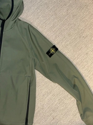 Grön Stone Island Junior jacka - Grön jacka från Stone Island Junior med svart dragkedja och ikonisk patch på vänster ärm. Jag klippte av den tvätt lappen men NFC-tagen är fortfarande kvar. Jackan har dessutom två framfickor med dragkedja och är tillverkad i ett unikt material. Passar lite mer åt till streetwear hållet.
