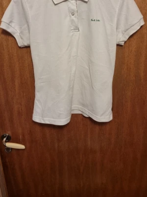 Vit pikétröja från Paul Smith - Klassisk vit pikétröja från Paul Smith med korta ärmar och krage. Tröjan har en liten grön broderad logga på bröstet och knappar vid halsen. Perfekt till både jeans och shorts för en clean look. Stl xl mer som en S