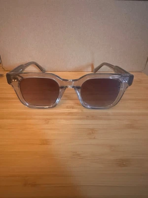  Chimi 04  - Snygga solglasögon från Chimi med transparent båge och glittriga silverdetaljer på skalmarna. Klassisk fyrkantig form som passar till många stilar. Mörka linser och trendig design – perfekta för att sticka ut i mängden.