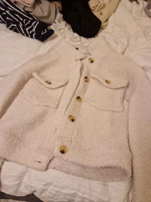 Beige teddyjacka med stora fickor - Säljer en beige teddyjacka med stora bröstfickor och guldiga knappar framtill. Jackan har en boxy passform och är gjord i mjukt, fluffigt teddy-material som ger en riktigt cozy vibe. Perfekt för dig som vill ha en trendig och bekväm jacka till hösten eller vintern. Passar s