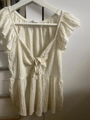 Vit volangblus med knyt fram - Supersöt vit blus med volangärmar och knyt framtill. Blusen har en lätt rynkad struktur och är tillverkad i ett tunt, luftigt material som passar perfekt till varma dagar. Den har en v-ringad hals och volangdetaljer längs ärmar och nederkant för en romantisk look.