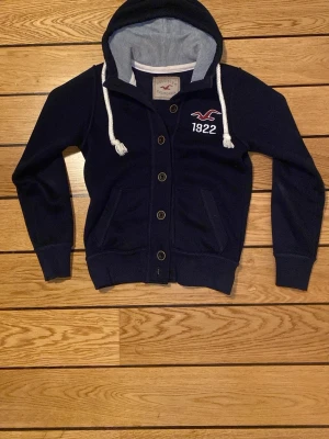 Mörkblå hoodie från Hollister - Mörkblå hoodie från Hollister med knappar framtill, vita dragsnören och broderad logga samt siffran 1922 på bröstet. Insidan av huvan är ljusgrå och materialet känns mjukt och bekvämt. Två fickor framtill och ribbade muddar vid ärmslut och nederkant.