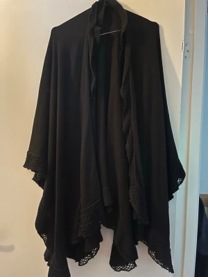 Svart sjal med virkade kanter - Stilren svart sjal med snygga virkade kanter och mjukt, följsamt tyg. Sjalen har en generös storlek och faller snyggt över axlarna. Perfekt att slänga över sig för en cool och avslappnad look.