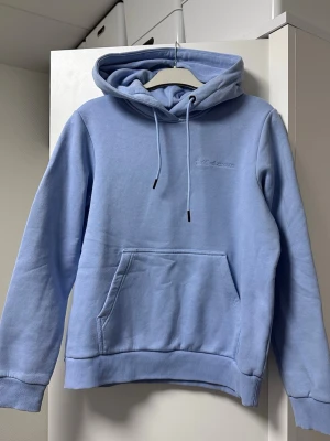 Ljusblå hoodie från Peak Performance - Snygg ljusblå hoodie från Peak Performance med broderad logga på bröstet. Klassisk känguruficka framtill, ribbade muddar och justerbar huva med dragsko. Perfekt för en chill och avslappnad stil. Tillverkad i mjukt bomullsmaterial.