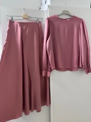 Rosa satinsätt med lång kjol och topp - Säljer ett snyggt tvådelat set i rosa satin, bestående av en lång, svepande kjol med omlott-design och en matchande långärmad topp med rund hals. Materialet är glansigt och mjukt, perfekt för dig som gillar stilrena och trendiga plagg.
