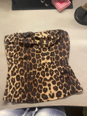 Leopardmönstrad tubtopp - Säljer en snygg tubtopp med leopardmönster i bruna och beige toner. Toppen är axelbandslös och har en mjuk, stretchig passform som sitter bekvämt. Perfekt för dig som vill sticka ut med ett djurmönster!