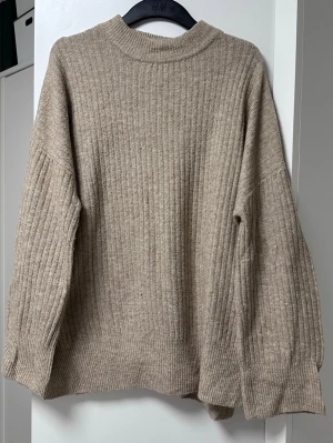 Beige ribbstickad tröja från H&M - En beige ribbstickad tröja från H&M med breda ärmar och hög krage. Tröjan har en avslappnad passform och är gjord i ett mjukt stickat material som passar perfekt för kyliga dagar. Enkel och stilren design som är lätt att matcha.