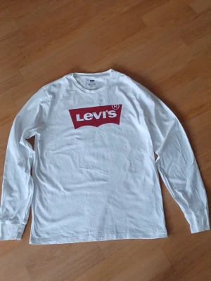 Vit långärmad Levi's t-shirt - Säljer en vit långärmad t-shirt från Levi's med det klassiska röda logotrycket på bröstet. Tröjan har rund hals och är gjord i mjuk bomull, perfekt för en avslappnad och stilren look.