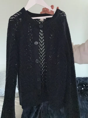 Svart virkad kofta från Gina Tricot - Snygg svart virkad kofta från Gina Tricot med långa ärmar och knappar framtill. Koftan har ett genomgående hålmönster som ger en trendig look och rund halsringning. Perfekt att slänga över en topp för en cool lager-på-lager-stil.