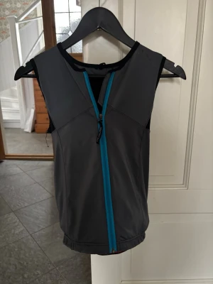 Svart/grå skyddsväst från Marker - Skyddsväst från Marker i svart och grått med blå dragkedja framtill. Västen är ärmlös och har en sportig design med elastisk nederkant och skyddande panel på ryggen. Perfekt för skidåkning eller andra actionsporter där extra ryggskydd behövs. Den har även band rundt midjan (karborre) den här använd som märks lite men inget som hindrar funktionen! Storleken ser ni på bilden