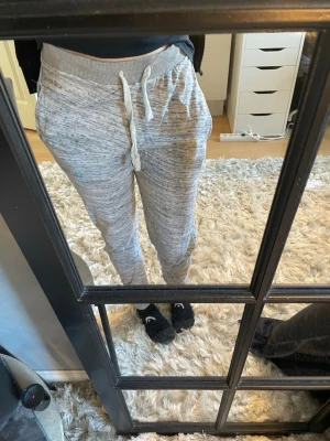 Gråmelerade mjukisbyxor med dragsko - Säljer ett par gråmelerade mjukisbyxor med bred resår i midjan och vit dragsko. Byxorna har muddar vid bensluten och två sidofickor. Perfekta för chill dagar hemma eller när du vill vara extra bekväm. Materialet är mjukt och stretchigt för maximal komfort. Väldigt tunnt material. Tror dom är köpta utomlands eller i stockholm för väldigt många år sedan