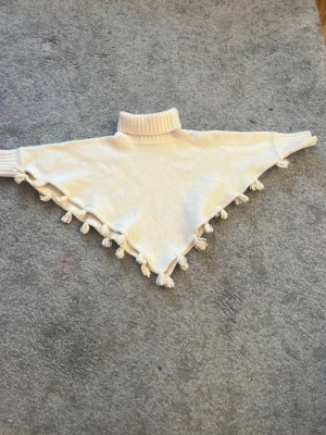 Vit stickad halsduk med tofsar - Superfin vit poncho-inspirerad tröja med polokrage och dekorativa tofsar längs kanterna. Tröjan har ribbade muddar vid hals och ärmslut och är perfekt för lager-på-lager-stil. Snygg och unik detalj till din garderob. 