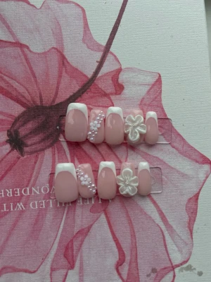 Press-on naglar med fransk och 3D-blomma - Snygga press-on naglar i ljusrosa med vit fransk tipp, dekorerade med vita pärlor och 3D-blommor. Naglarna har en kort, fyrkantig form och ger en söt och trendig look. Perfekt för dig som vill ha en unik och kreativ nageldesign.