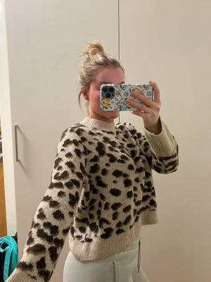 Beige leopardmönstrad stickad tröja - Säljer en beige stickad tröja med leopardmönster i svart och brunt. Tröjan har rund halsringning, långa ärmar och ribbade muddar vid ärmslut och nederkant. Perfekt för dig som gillar djurmönster och vill ha en trendig look.
