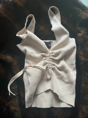 Beige ribbad linnetopp med snörning - Trendig beige linnetopp från Design Studio med ribbad struktur och snygg snörning framtill. Toppen är ärmlös med breda axelband och croppad passform, perfekt för att skapa en cool och avslappnad look.