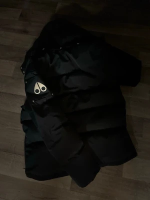 Svart pufferjacka med pälsluva - Säljer en svart pufferjacka från Moose Knuckles med pälsluva och silvriga detaljer. Jackan har dragkedja, knappar och två fickor framtill samt en logga på ärmen. Perfekt för kalla vinterdagar och riktigt snygg streetstil.