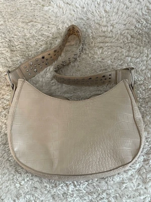 Beige axelväska med nitar och crocomönster - Snygg beige axelväska med crocomönster och bred axelrem dekorerad med silvriga nitar. Väskan har rundad form och dragkedja upptill. 