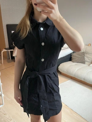 Svart denimklänning med knytskärp - Kort svart skjortklänning med stora vita knappar framtill och knytskärp i midjan. Klänningen har korta ärmar och klassisk krage, perfekt för en trendig och stilren look.