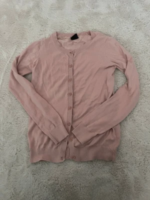 Ljusrosa cardigan med knappar - Söt ljusrosa cardigan med rund halsringning och knäppning framtill. Långa ärmar och ribbade muddar vid ärmslut och nederkant. Perfekt att slänga över en topp för en chill look.