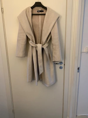Beige kappa med huva från Zara - Stilren beige kappa från Zara med stor huva och knytskärp i midjan. Kappan är lång, har raka ärmar och är tillverkad i ett mjukt tyg som ger en lyxig känsla. Perfekt för kyliga dagar när du vill vara både varm och snygg.