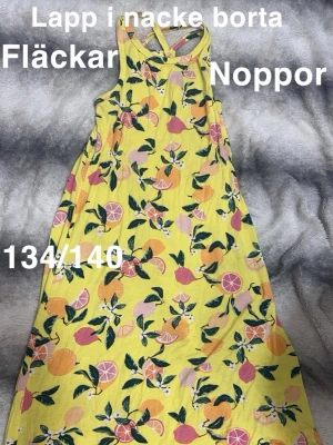 Gul sommarklänning med citrusmönster - Snygg gul långklänning med färgglatt citrusmönster i rosa, orange och grönt. Klänningen har smala axelband som korsas i ryggen. perfekt för varma dagar. Fräsch och lekfull stil som sticker ut.🌸Lappen i nacken är borta. Noppor. Fläckar. Storlek 134/140