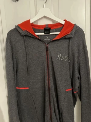 Grå dress från Hugo Boss. - Snygg grå dress från Hugo Boss med dragkedja, huva och röda detaljer på insidan av huvan och vid fickorna. Märkets logga syns framtill. Tillverkad i mjukt material som känns skönt mot huden. Perfekt för en avslappnad och trendig look. Hoodien är i M och Byxorna S. 