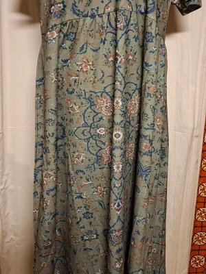 Grön långklänning med blommigt mönster - Långklänning i mjukt grönt tyg med blommigt mönster i blått, beige och vitt. Klänningen har korta ärmar och en avslappnad passform med markerad midja. Perfekt för dig som gillar vintageinspirerad stil och unika detaljer.