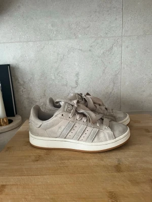 Beige/ljus rosa Adidas Campus - Säljer ett par beige/ ljus rosa Adidas campus i mocka med klassiska tre ränder på sidan och breda matchande skosnören. Skorna har låg profil och vit sula, samt Adidas-logga på plösen. Perfekta för dig som gillar stilrena och neutrala färger.
