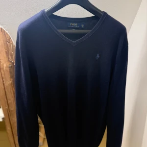 Mörkblå V-ringad tröja Polo Ralph Lauren - Klassisk mörkblå V-ringad tröja från Polo Ralph Lauren med diskret broderad logga på bröstet. Tröjan har en stilren design och är tillverkad i mjukt material som passar perfekt till både jeans och skjorta.