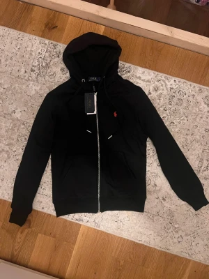 Svart hoodie från Polo Ralph Lauren - Svart hoodie med dragkedja från Polo Ralph Lauren. Klassisk röd logga broderad på bröstet, två fickor framtill och justerbar huva med snören. Tillverkad i mjukt bomullsmaterial för en skön och avslappnad look.