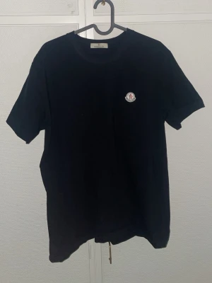 Svart Moncler t-shirt med logotyp - Säljer en svart t-shirt från Moncler med klassisk rund hals och korta ärmar. På bröstet finns Monclers ikoniska logotyp. T-shirten är enkel och stilren, perfekt för dig som gillar exklusiva märken och minimalistisk design. Sitter som M
