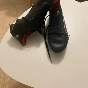 Svarta Puma King fotbollsskor ag/fg - Säljer ett par svarta Puma King fotbollsskor med orangea och vita detaljer. Skorna har klassisk snörning, låg profil och slitstark ovandel i syntetmaterial. Perfekta för dig som vill ha bra touch och kontroll på planen. Jag har bara använt dom typ 5 gånger eftersom de blev försmå under en skada.