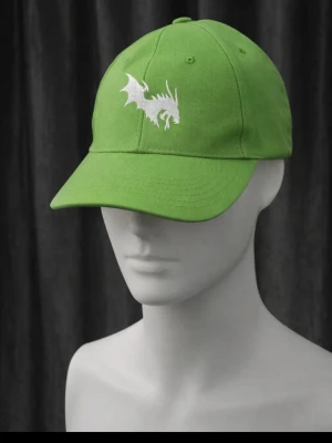 Grön närcon keps med drakbrodyr - Snygg grön keps med vit broderad drake framtill. Klassisk böjd skärm och justerbar baksida. Perfekt accessoar för att sticka ut och ge outfiten en cool touch.