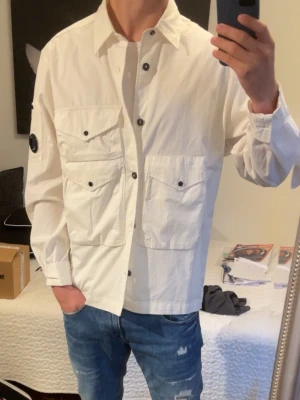 Vit overshirt från C.P. Company - Stilren vit overshirt från C.P. Company med fyra stora fickor framtil. Overshirten har knappstängning och är gjord i ett lätt bomullsmaterial. Perfekt för lager-på-lager och ger en clean look. Den är knappt använd och har inga defekter! Är öppen för förhandling om de vore så.