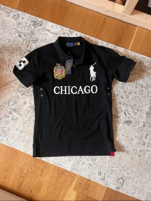 Svart Ralph Lauren polo med CHICAGO-tryck - Snygg svart pikétröja från Ralph Lauren med stort vitt CHICAGO-tryck på bröstet, broderad logga och emblem. Klassisk krage med knappar och korta ärmar. Tillverkad i mjuk bomull för skön känsla och stilren look.