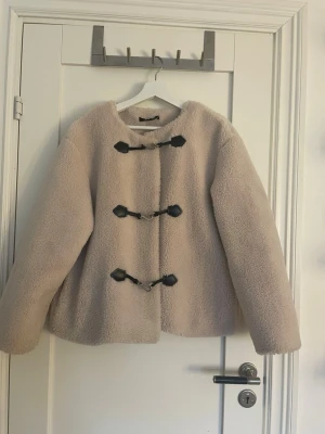 Beige teddyjacka med svarta detaljer - Mysig beige teddyjacka med tre svarta toggle-knäppen framtill. Materialet är fluffigt och ger en trendig look. Perfekt för dig som vill ha en varm och stylish jacka till hösten eller våren.