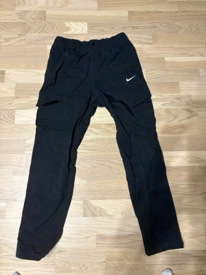 Svarta cargobyxor från Nike - Säljer ett par svarta cargobyxor från Nike med klassisk vit swoosh-logga på benet. Byxorna har flera praktiska fickor och elastisk midja för skön passform. Perfekta för en sportig och avslappnad stil. Materialet känns lätt och smidigt.