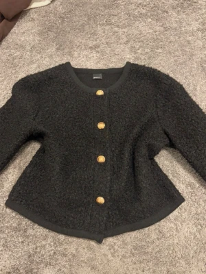Svart bouclé cardigan med guldknappar - Svart cardiganjacka från Gina tricot i bouclé-material med rund halsringning och fyra dekorativa guldknappar framtill. Jackan har lång ärm och en något boxig, kortare passform. Perfekt för att addera lite lyxig känsla till din outfit. Använd 1 gång, lite nopprig i materialet 
