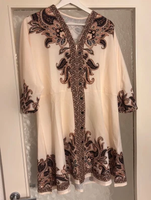 Vit och brun mönstrad klänning - Unik vit klänning med bruna och svarta broderade detaljer och v-ringning. Klänningen har trekvartsärm och knytband i nacken. Lätt utsvängd modell med dekorativa mönster längs kanter och ärmslut. Perfekt för dig som gillar bohemisk stil.