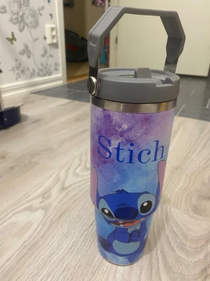 Stitch vattenflaska med handtag - Säljer en vattenflaska i rostfritt stål med motiv av Stitch från Disney. Flaskan har ett grått lock med praktiskt bärhandtag och färgglad blå-lila bakgrund. Perfekt för att ta med dryck till skolan eller träningen.