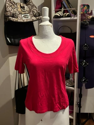 Betty Barclay Topp - Elegant topp från Betty Barclay i en stark röd/fuchsia färg. Modellen har en klassisk rund halsringning med en diskret dekorativ detalj runt kanten som ger ett stilrent uttryck. Det mjuka och stretchiga tyget gör toppen bekväm att bära och enkel att styla både till vardags och mer uppklädda outfits.
