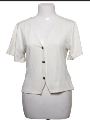 Vit kortärmad blus med knappar - Stilren vit blus med V-ringning och tre svarta knappar framtill. Blusen har korta ärmar och en avslappnad passform, perfekt för en enkel och fräsch look. Tillverkad i ett mjukt, lätt material som känns skönt mot huden.