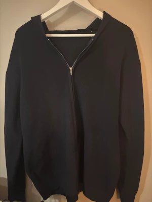 Svart hoodie med dragkedja - Mysig svart hoodie med dragkedja framtill och huva. Tillverkad i ett stickat material som ger en avslappnad och stilren look. Perfekt för att slänga på sig över en t-shirt. Långa ärmar och ribbade muddar.