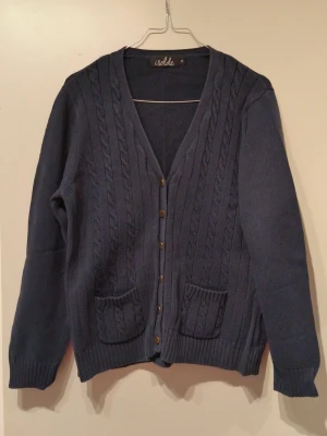 Mörkblå kabelstickad kofta Isolde - Mysig mörkblå kofta från Isolde med kabelstickat mönster, v-ringning och knappar framtill. Två praktiska fickor framtill och ribbade muddar vid ärmslut och nederkant. Perfekt för lager på lager och en chill look. Knapparna är lite missfärgade annars i väldigt bra skick.