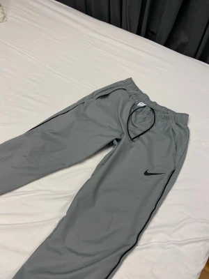 Nike dri fit byxor - Snygga gråa mjukisbyxor från Nike med svart logga på vänster ben. Byxorna har resår i midjan med dragsko och sidofickor. Perfekta för chill dagar eller träning. Tillverkade i mjukt och bekvämt material.