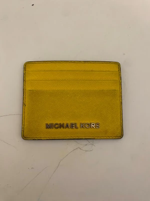 Gul korthållare från Michael Kors - Snygg gul korthållare från Michael Kors i tåligt läder. Framsidan har plats för flera kort och pryds av en metallogo. Baksidan har ett genomskinligt fack perfekt för ID eller busskort. Enkel och stilren design som sticker ut.