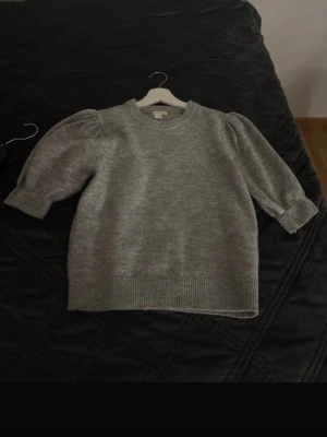 Grå stickad tröja från Gina Tricot - Mysig grå stickad tröja från Gina Tricot med puffärmar och ribbade muddar vid hals, ärmslut och nederkant. Tröjan har en avslappnad passform och är perfekt för lager-på-lager-stil. Enkel att matcha med jeans eller kjol.