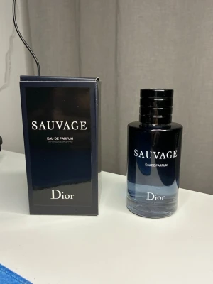 Dior Sauvage Eau de Parfum - Dior Sauvage Eau de Parfum med elegant mörkblå glasflaska och matchande svart kartong. Flaskan har en stilren, rund form med silverfärgad text och en svart kork med Dior-loggan på toppen. En ikonisk doft som utstrålar lyx och modernitet.