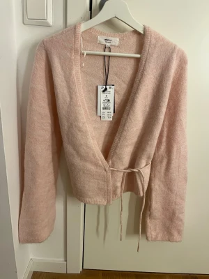 Ljusrosa omlottkofta från Gina Tricot - Superfin ljusrosa stickad omlottkofta från Gina Tricot med knytband i midjan och djup v-ringning. Koftan har långa ärmar och är gjord i ett mjukt, fluffigt material som känns lyxigt och trendigt. Aldrig använd med prislappen kvar - originalpris: 399 kr