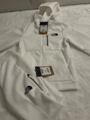 Vit hoodie och byxa från The North Face - Säljer ett set med vit hoodie och matchande byxa från The North Face. Hoodien har dragkedja, fickor och logotyp på bröstet. Byxan har elastisk midja, dragkedja vid fickan och logotyp på benet. Perfekt för en avslappnad och sportig look.